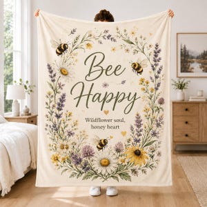 Peut inclure: Une couverture en polaire crème ornée d'une couronne florale de marguerites, de lavande et de fleurs sauvages avec trois abeilles. Le texte Bee Happy et Wildflower soul, honey heart est écrit en brun foncé au centre du tissu.
