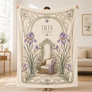 May include: A cream-colored throw blanket featuring an Art Nouveau-style illustration of purple iris flowers surrounding a garden archway and a wicker chair. The text IRIS GARDEN is printed in the center. The design includes intricate green vines.