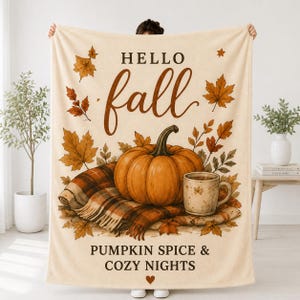 Könnte beinhalten: Eine cremefarbene Wohndecke mit dem Text HELLO fall und PUMPKIN SPICE & COZY NIGHTS. Das Design zeigt einen großen orangefarbenen Kürbis, eine karierte Decke, eine Keramiktasse und verstreute Herbstblätter in Braun- und Orangetönen.