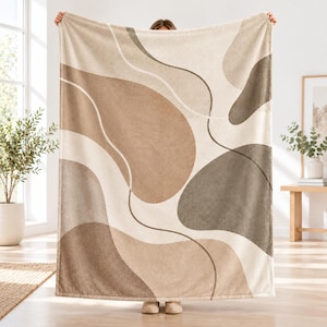 May include: A rectangular throw blanket featuring a large-scale abstract pattern of organic, fluid shapes in varying shades of beige, tan, taupe, and dark brown, separated by thin, curved lines against a light cream-colored background.