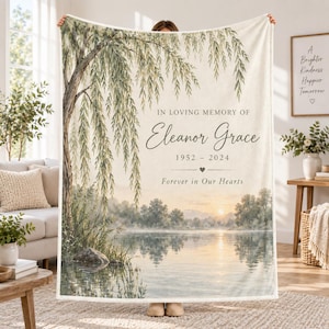 May include: A cream-colored memorial throw blanket featuring a weeping willow tree over a calm lake at sunset. The text reads: IN LOVING MEMORY OF Eleanor Grace 1952 - 2024 Forever in Our Hearts. The design includes a small heart icon below the dates.