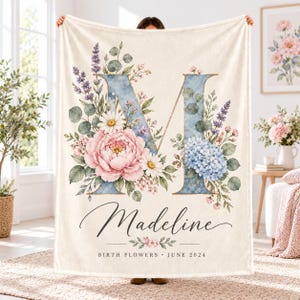 May include: A cream-colored fleece blanket featuring a large blue letter M decorated with watercolor-style pink peonies, blue hydrangeas, daisies, and lavender. The name Madeline is printed in black script below, with the text BIRTH FLOWERS JUNE 2024.