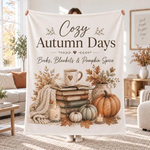 May include: A cream-colored throw blanket featuring the text "Cozy Autumn Days" and "Books, Blankets & Pumpkin Spice." The design includes a stack of books, a mug with a heart, a pumpkin, a gourd, two candles, and a knitted throw blanket.