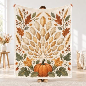 May include: A cream-colored throw blanket featuring a central orange pumpkin at the bottom, surrounded by green leaves and yellow flowers. Above the pumpkin, a fan-shaped arrangement of light brown pumpkin seeds is flanked by wheat stalks.