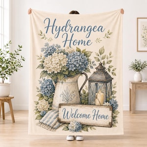 Può includere: Una coperta color crema con una composizione floreale di ortensie blu e bianche in una brocca, una lanterna con una candela e un cartello in legno con il testo Hydrangea Home e Welcome Home in caratteri blu.