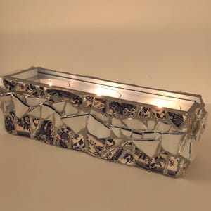 May include: A rectangular candle holder featuring a mosaic exterior made of broken white and blue floral patterned ceramic shards. The long, narrow vessel contains three lit tea light candles, with the shards held together by silver-toned grout.