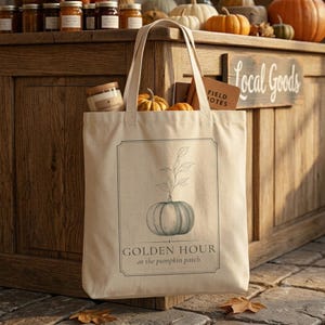 May include: A beige canvas tote bag featuring a dark green line drawing of a pumpkin with leaves. The text reads GOLDEN HOUR at the pumpkin patch. The bag contains a glass jar with a tan lid and a brown notebook, with a small pumpkin visible.