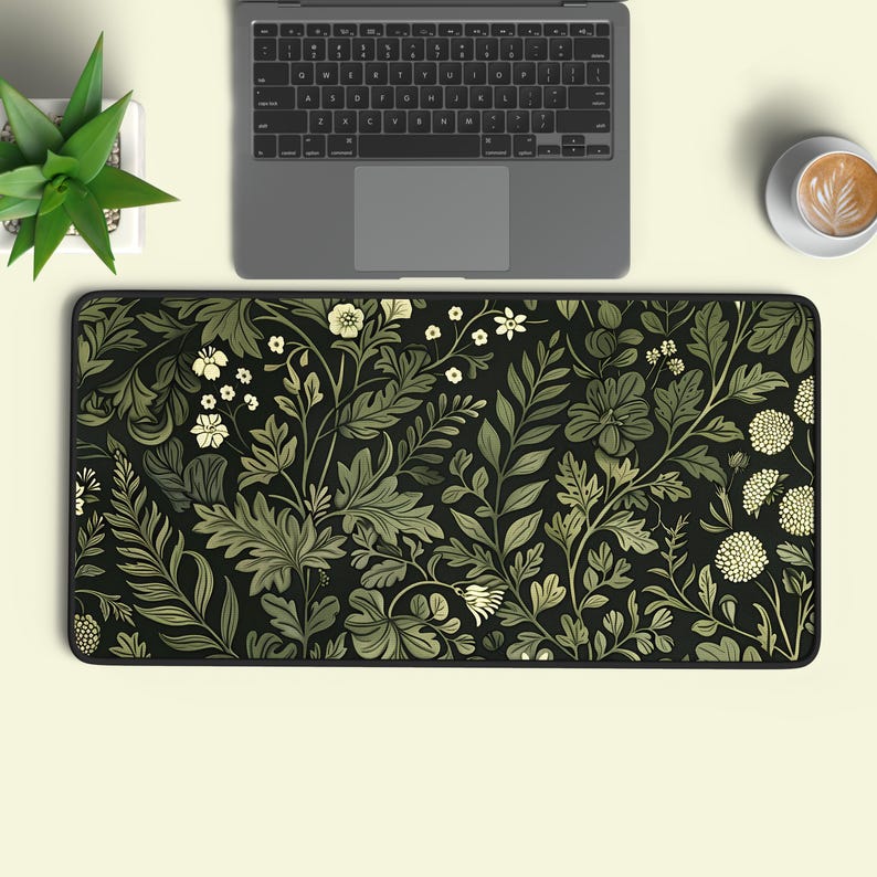 May include: A rectangular desk mat featuring a black background with a dense, repeating botanical pattern of light green and cream-coloured leaves, ferns, and various small flowers, finished with a black stitched edge along the entire perimeter.