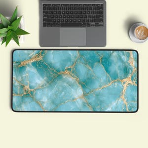 May include: A rectangular desk mat featuring a vibrant turquoise and teal marble pattern with intricate gold veining throughout. The mat has a solid black border and is positioned below a grey laptop, a small green succulent, and a white coffee mug.