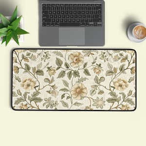 May include: A rectangular desk mat featuring a repeating floral pattern with beige flowers and muted green leaves on a cream-coloured background. The mat has a black stitched border and is positioned below a laptop, a small potted plant, and a coffee cup.