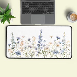 May include: A rectangular desk mat featuring a watercolour-style print of wildflowers in shades of blue, lavender, and soft peach against a white background. The mat has a black border and is positioned below a laptop, a small potted plant, and a coffee cup.