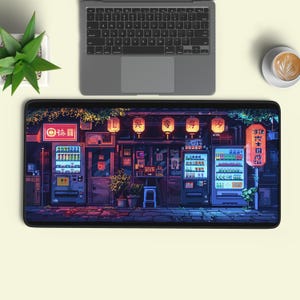Può includere: Un tappetino da scrivania rettangolare con un'illustrazione in pixel art di una scena stradale notturna con lanterne luminose, due distributori automatici e una vetrina. L'opera mostra vivaci tonalità di viola, blu e arancione.