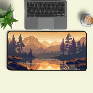 May include: A rectangular desk mat featuring a landscape illustration of a mountain range, pine trees, and a calm lake at sunset. The scene uses a warm colour palette of orange, gold, and deep blue, with the mountains reflected in the water.