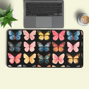 May include: A rectangular desk mat featuring a repeating pattern of watercolour-style butterflies in shades of blue, pink, yellow, and orange set against a solid black background. The mat is positioned below a laptop, a small potted plant, and a coffee cup.