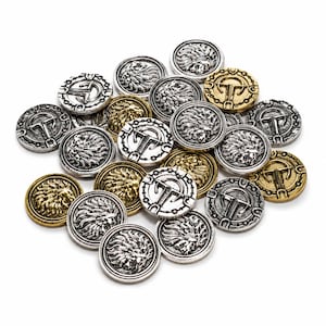 Puede incluir: Una colección de monedas metálicas circulares en tonos plateados y dorados. Cada moneda presenta un relieve detallado de una cabeza de león o un símbolo estilizado en forma de T rodeado por un borde decorativo. Las monedas están amontonadas.