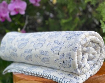 Könnte beinhalten: Eine weiße Baumwolldecke mit einem blau-weißen Muster aus stilisierten Schiffen. Die Decke ist aufgerollt und steht auf einem Holzhocker.