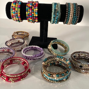 May include: A display of colorful beaded bracelets, some with turquoise, gold, and brown beads, others with purple, pink, and white beads. The bracelets are arranged on a black stand.