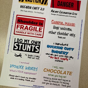 May include: A sheet of colorful name badges with humorous text and illustrations. The badges feature phrases like "Rotator Cuff 2.0", "Shoulder is Fragile", "I Do My Own Stunts", "I Survived Shoulder Surgery", and "I Will Need Chocolate".