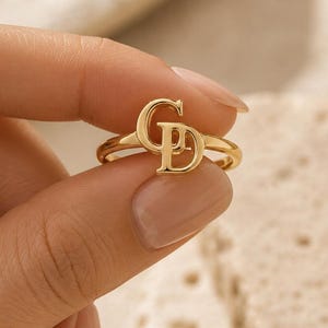 May include: A gold ring featuring a polished finish and a prominent, stylized monogram centerpiece consisting of the uppercase letters G and D intertwined. The band is thin and circular, showcasing a smooth, reflective metallic surface throughout.