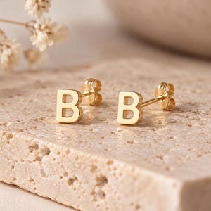 May include: A pair of gold stud earrings, each featuring the capital letter B in a block font. The earrings have a polished finish and include matching gold butterfly-style earring backs, presented as a set of two individual letter pieces.