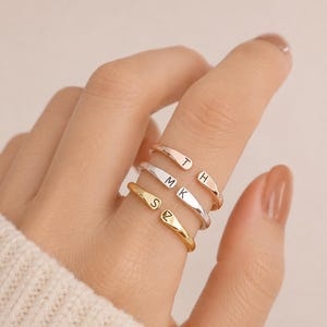 Könnte beinhalten: Drei offene Ringe in Roségold-, Silber- und Goldtönen. Jeder Ring hat zwei flache, abgerundete Enden, die mit einzelnen Großbuchstaben graviert sind. Der roségoldene Ring zeigt T und H, der silberne Ring M und K und der goldene Ring S und Z.