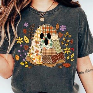 May include: A charcoal grey short-sleeve t-shirt featuring a central graphic of a ghost composed of various autumn-themed fabric patches. The ghost design includes plaid, floral, and pumpkin patterns surrounded by colourful leaves and small flowers.