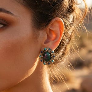 Pode incluir: Um brinco oval com uma pedra central escura e iridescente rodeada por uma moldura de metal dourado texturizado. A borda externa é acentuada com pequenas contas redondas turquesa e azul-escuras dispostas em um círculo.