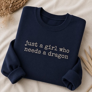 Könnte beinhalten: Ein marineblaues Sweatshirt mit Rundhalsausschnitt und dem gestickten Text "just a girl who needs a dragon" in hellbeigem Garn. Das Kleidungsstück hat lange Ärmel mit gerippten Bündchen und einen gerippten Halsausschnitt, flach zusammengelegt.