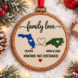May include: A round wooden embroidery hoop ornament featuring the embroidered text family love, a blue map of Florida, a green map of North Carolina, and the text OLIVIA, MOM & DAD, and KNOWS NO DISTANCE with a small red heart.