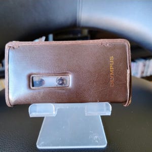Op de afbeelding: Een rechthoekige bruine lederen etui met een getextureerd oppervlak en het woord OLYMPUS in gouden letters aan de rechterkant. De etui heeft een horizontale rechthoekige uitsparing die een deel van een cassettebandje aan de binnenkant toont.