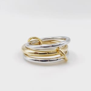 May include: A multi-band ring featuring interlocking circular bands in alternating polished gold and silver tones. The design consists of several smooth, rounded metal loops that are connected and layered to create a single cohesive piece of jewelry.
