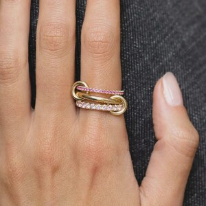 May include: A gold ring featuring three interconnected bands. The top band is set with small pink stones, the middle band is a plain polished gold link, and the bottom band is set with small clear white stones, creating a layered appearance.