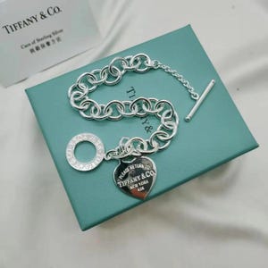Può includere: Un bracciale in argento con chiusura a barra a T e un ciondolo a forma di cuore inciso con il testo Please Return To Tiffany & Co. New York 925. Il bracciale presenta un'etichetta circolare e poggia su una scatola quadrata color ottanio.