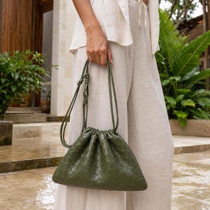 Puede incluir: Un bolso tipo sobre de color verde oliva con un exterior de cuero trenzado, cierre de cordón fruncido en la parte superior y una correa de hombro fina y ajustable. El bolso tiene una forma redondeada suave y un acabado liso y uniforme.