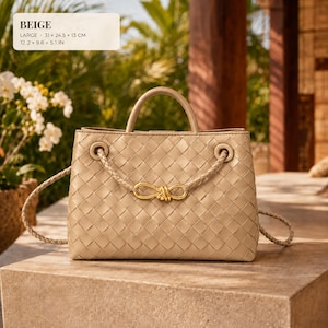 K&ouml;nnte beinhalten: Eine beige geflochtene Ledertasche mit einem strukturierten Tragegriff und einem geflochtenen Schulterriemen mit einer goldfarbenen Metallschleife. Die Tasche misst 31 x 24,5 x 13 cm, wie im Text auf dem Bild angegeben.
