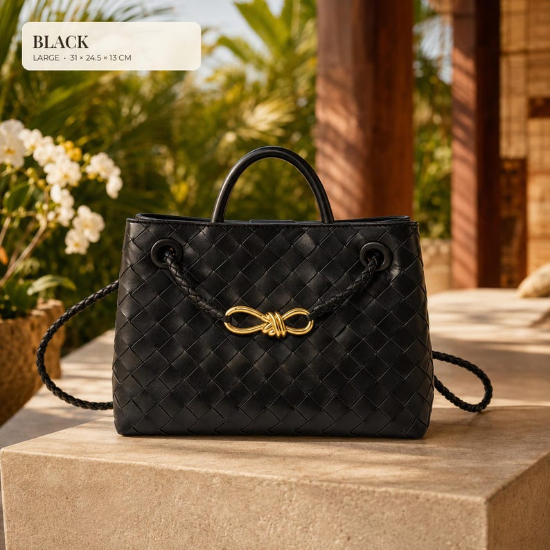 K&ouml;nnte beinhalten: Eine schwarze Handtasche mit geflochtener Lederstruktur, ausgestattet mit einem Tragegriff und einem geflochtenen Schulterriemen. Ein goldfarbenes Metallornament in Knotenform ist vorne mittig angebracht. Text: BLACK LARGE 31 x 24.5 x 13 CM.