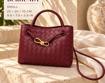 Könnte beinhalten: Eine kleine bordeauxrote Handtasche mit geflochtenem Leder, einem Tragegriff und einem geflochtenen Schulterriemen. Die Tasche hat ein goldenes Metallknotendetail auf der Vorderseite. Der Text lautet: BURGUNDY, SMALL, 25 x 20 x 10 CM, 9.8 x 7.9 x 3.9 IN.