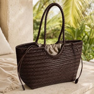 May include: A dark brown woven tote bag featuring a herringbone pattern, two braided shoulder straps, and a beige drawstring fabric interior closure. The bag has a curved top opening and long, thin side cords with small rounded end caps.