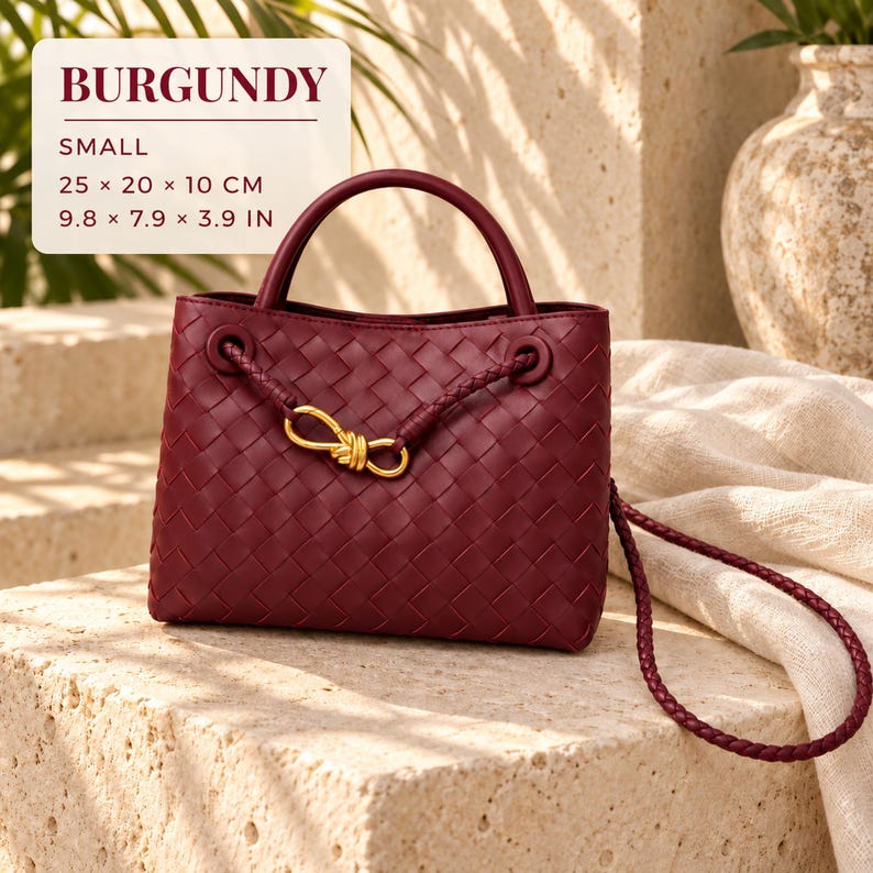 K&ouml;nnte beinhalten: Eine kleine bordeauxrote Handtasche mit geflochtenem Leder, einem Tragegriff und einem geflochtenen Schulterriemen. Die Tasche hat ein goldenes Metallknotendetail auf der Vorderseite. Der Text lautet: BURGUNDY, SMALL, 25 x 20 x 10 CM, 9.8 x 7.9 x 3.9 IN.