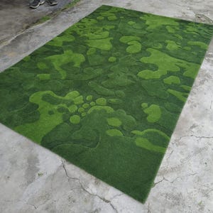 May include: A rectangular area rug featuring a textured, multi-tonal green surface with an abstract, organic pattern of raised and recessed shapes that mimic the appearance of moss or foliage, creating a layered, three-dimensional effect.