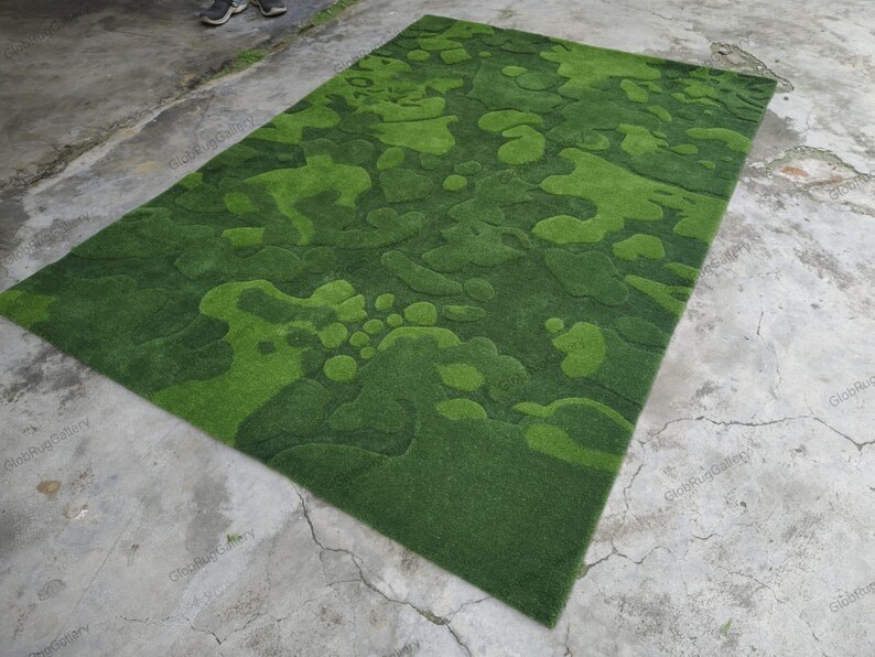 May include: A rectangular area rug featuring a textured, multi-tonal green surface with an abstract, organic pattern of raised and recessed shapes that mimic the appearance of moss or foliage, creating a layered, three-dimensional effect.