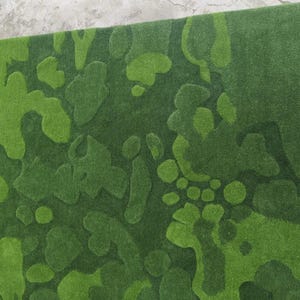 May include: A rectangular area rug featuring a textured, abstract camouflage pattern in various shades of green. The design consists of organic, rounded shapes in light, medium, and dark green tones, creating a layered, three-dimensional effect.