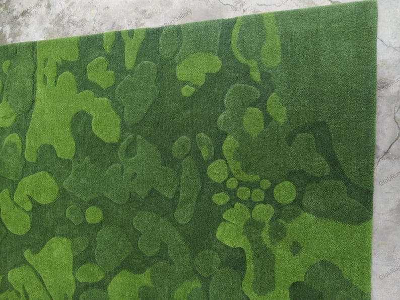 May include: A rectangular area rug featuring a textured, abstract camouflage pattern in various shades of green. The design consists of organic, rounded shapes in light, medium, and dark green tones, creating a layered, three-dimensional effect.