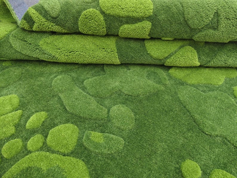 May include: A plush area rug featuring a textured, tufted surface with an organic, irregular pattern of overlapping shapes in varying shades of lime green and forest green. The rug is folded over, revealing a dense, soft pile and a consistent color palette.