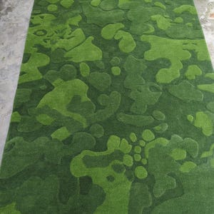 May include: A rectangular area rug featuring a textured, abstract camouflage pattern in various shades of green. The design consists of organic, rounded shapes with varying pile heights that create a three-dimensional effect across the surface.
