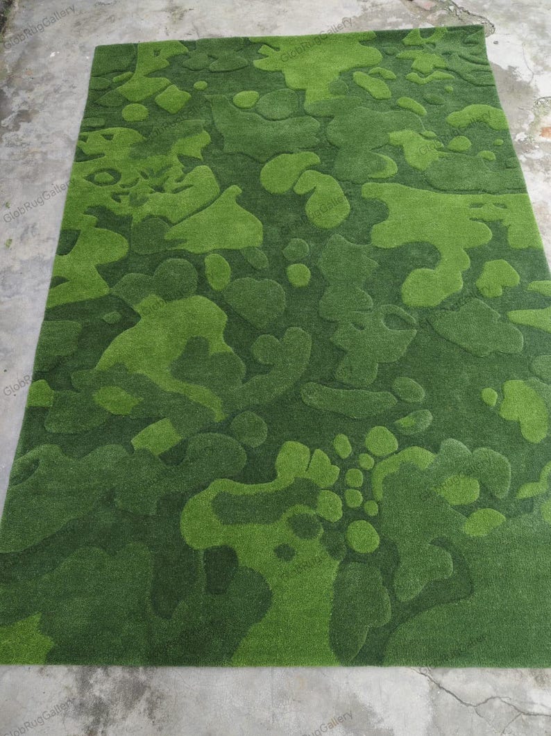 May include: A rectangular area rug featuring a textured, abstract camouflage pattern in various shades of green. The design consists of organic, rounded shapes with varying pile heights that create a three-dimensional effect across the surface.