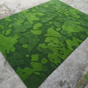 May include: A rectangular area rug featuring a textured, carved surface with an abstract camouflage pattern in various shades of green. The design consists of organic, irregular shapes in light lime and deep forest green tones with pile height.