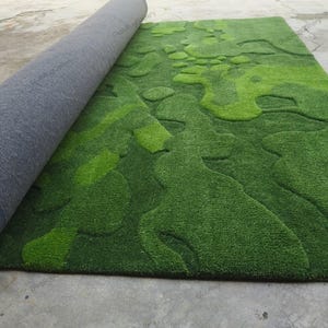May include: A rectangular area rug featuring a textured, multi-tonal green surface with an abstract, organic pattern of raised and recessed shapes. The rug is partially rolled, revealing a dark gray, felt-like backing on the underside.
