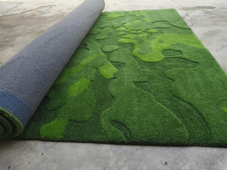 May include: A rectangular area rug featuring a textured, multi-tonal green surface with an abstract, organic pattern of raised and recessed shapes. The rug is partially rolled, revealing a dark gray, felt-like backing on the underside.