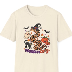 May include: A cream-colored short-sleeve t-shirt featuring a pixel-art style graphic of a snake wearing a witch hat, a ghost, a black cat, pumpkins, bats, and candy corn. The text "ssssssscary" is printed in purple below the snake illustration.